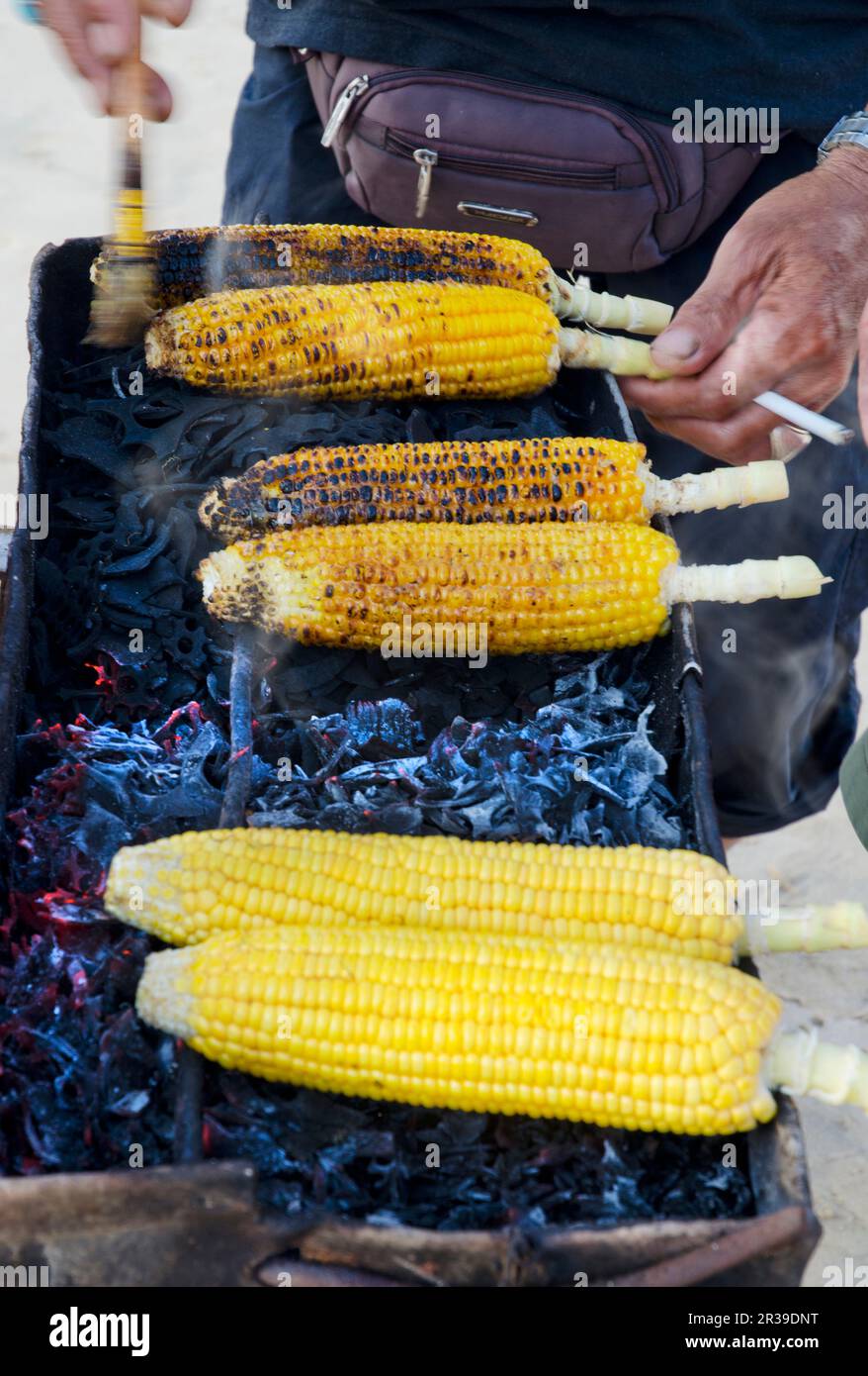 Un vendeur de rue balinais vendant des épis de maïs épicés sur un barbecue au charbon de bois avec une cigarette dans la main Banque D'Images
