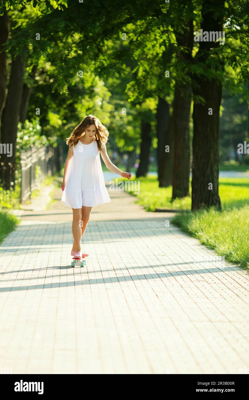 Belle femme urbaine en robe blanche avec un skate rose. Jeune fille à cheval dans le parc sur un skateboard Banque D'Images