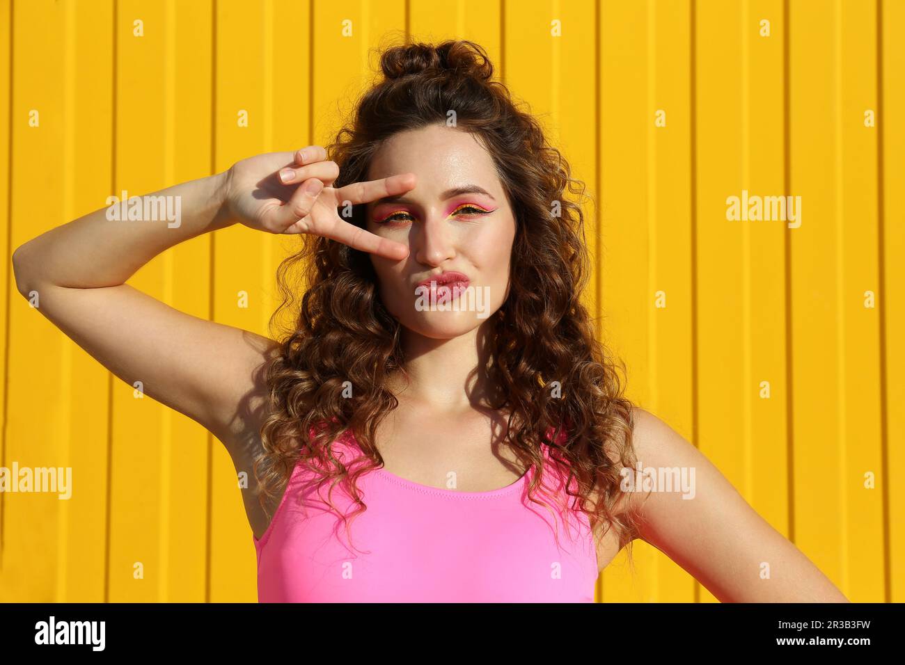 Femme faisant signe de la paix geste près de l'oeil devant le mur jaune Banque D'Images