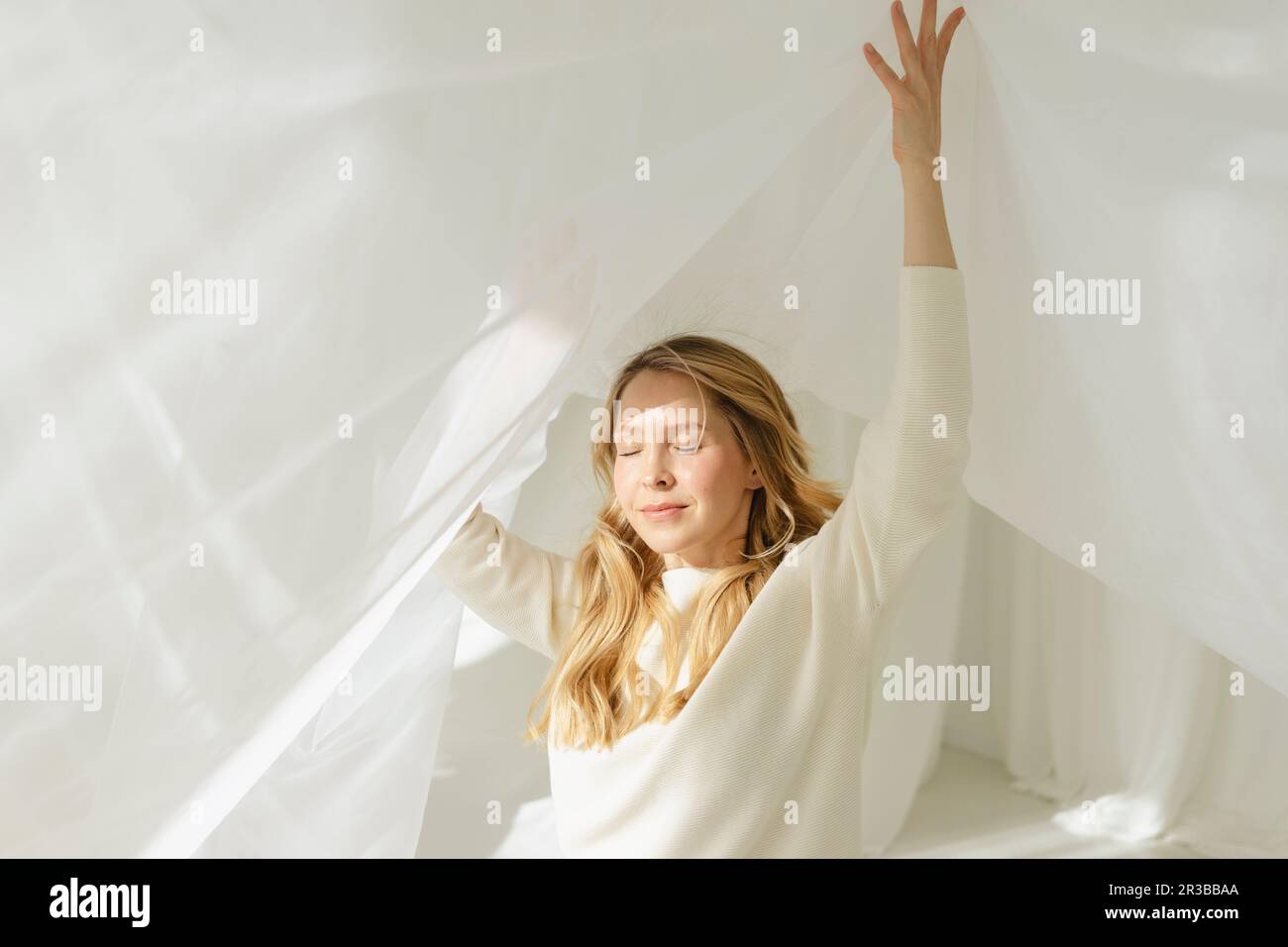 Femme blonde avec les yeux fermés et les bras levés sous un rideau blanc translucide Banque D'Images