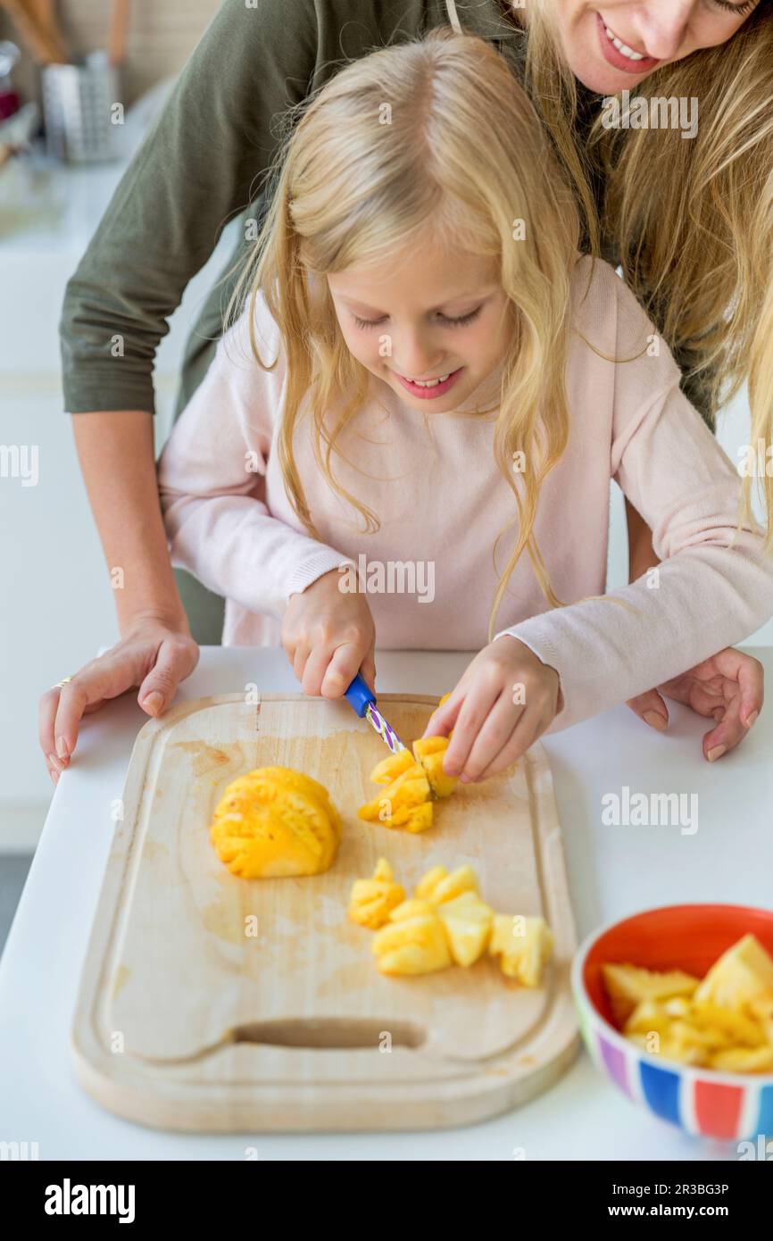 Fille souriante apprenant à hacher des ananas sur la planche à découper par la mère à la maison Banque D'Images