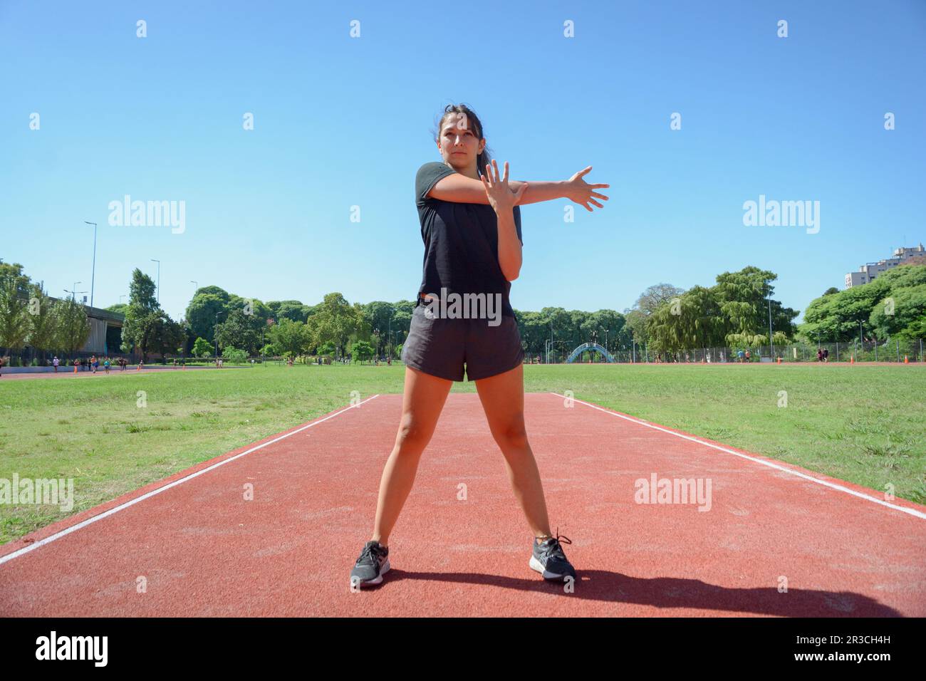vue avant de la jeune femme latine s'entraîner sur le circuit sportif, faire des étirements de bras, se tenir sur la piste se réchauffer en préparation pour courir. Banque D'Images