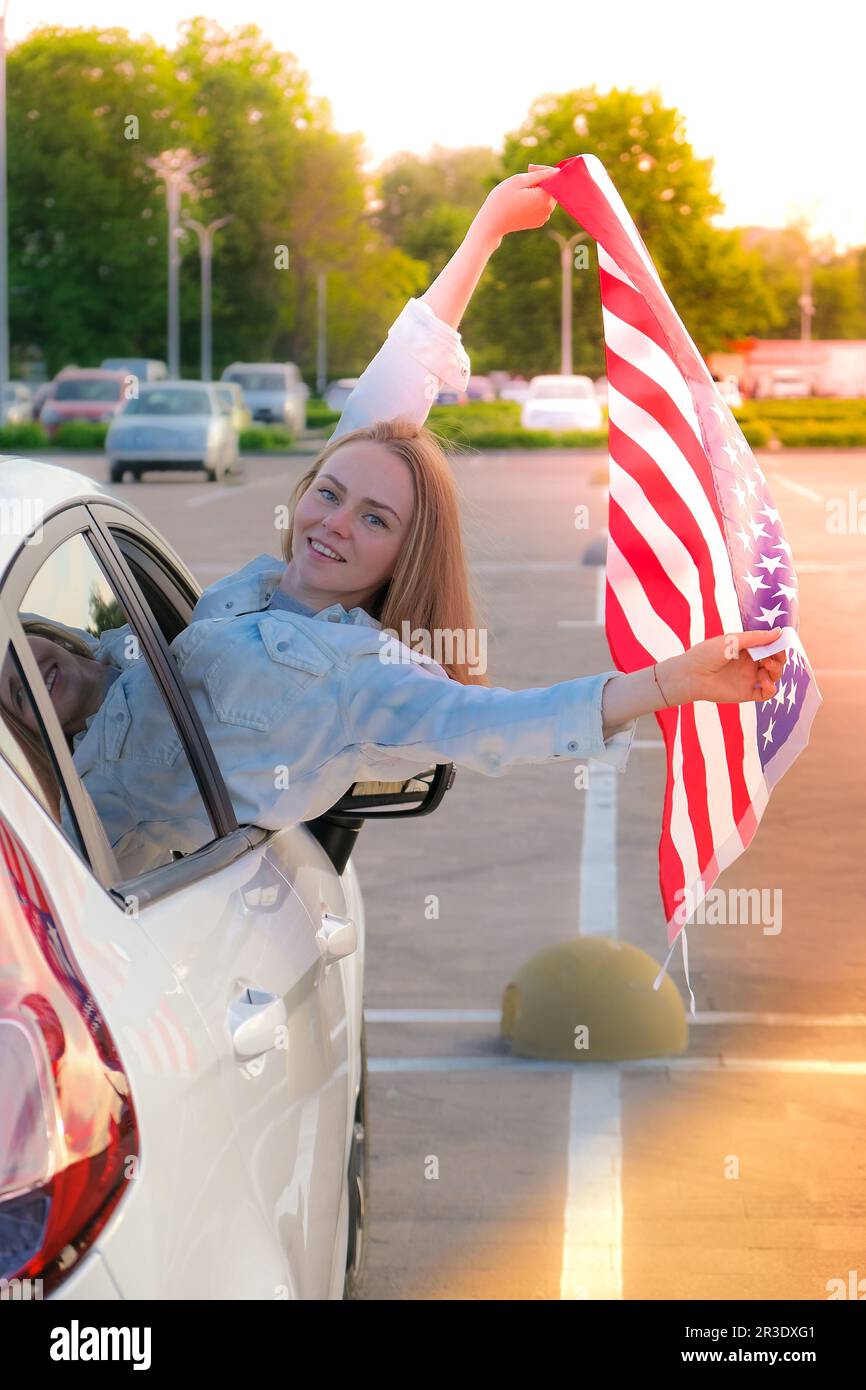 Jeune femme blonde millénaire à la recherche d'une voiture avec drapeau américain. Drapeau des états-unis entre ses mains. Indépendance de 4 juillet Banque D'Images