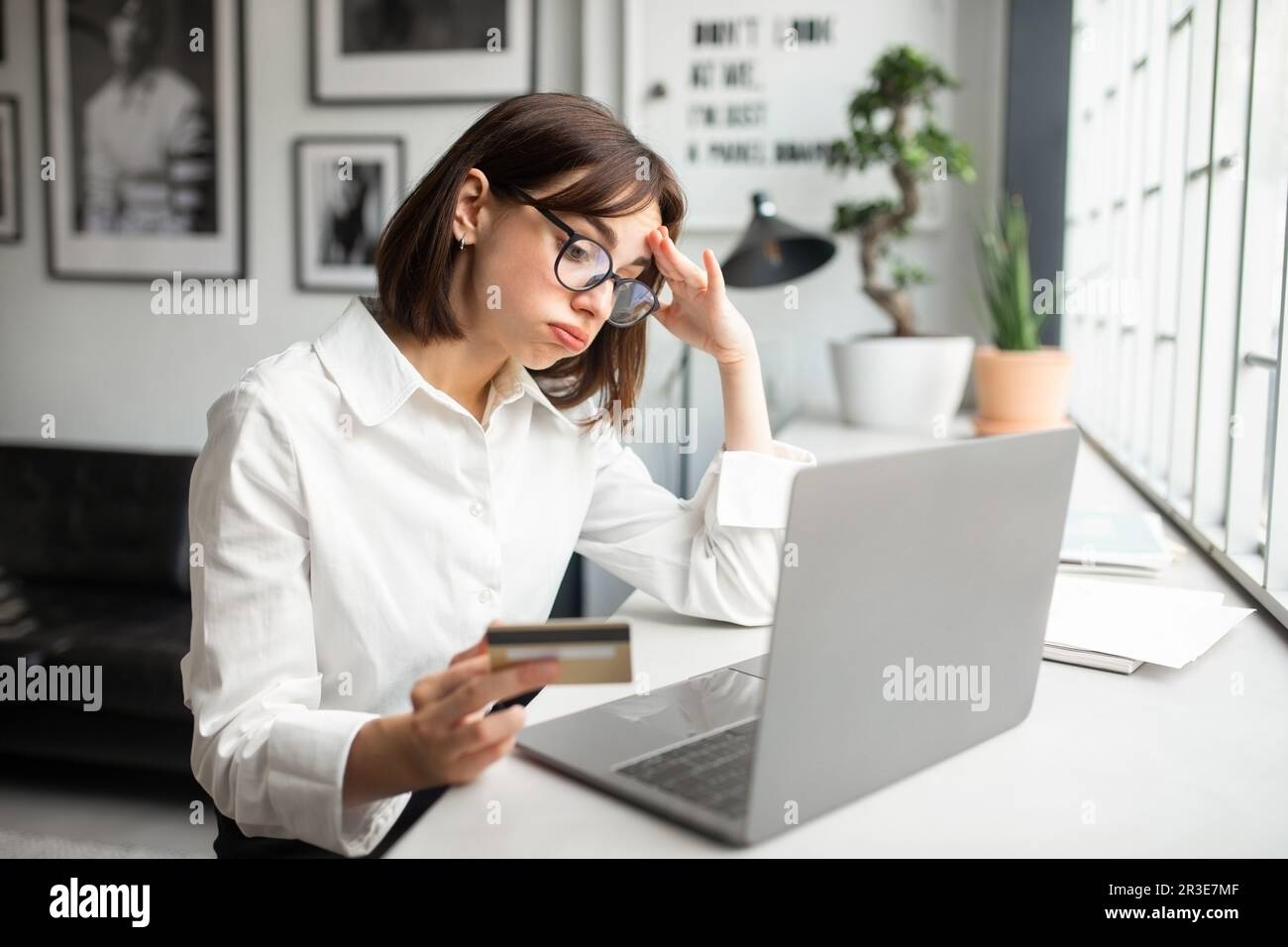 Déçue jeune femme d'affaires travaillant dans un bureau ou un café confortable, tenant une carte de crédit et essayant de faire l'achat Banque D'Images
