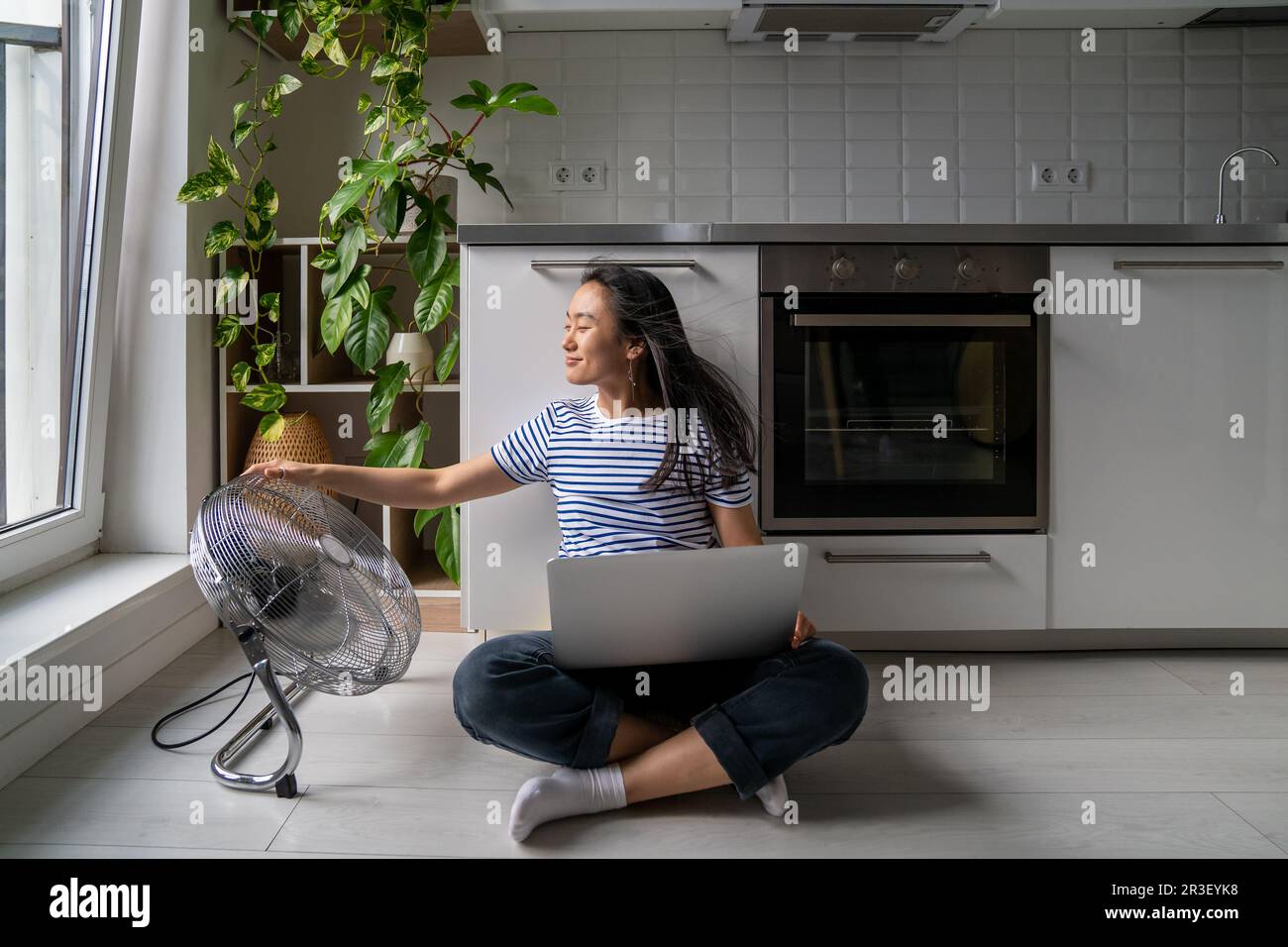 Femme asiatique confortable allumez le ventilateur de plancher tout en travaillant avec un ordinateur portable joyeuse auto-économie de la chaleur Banque D'Images
