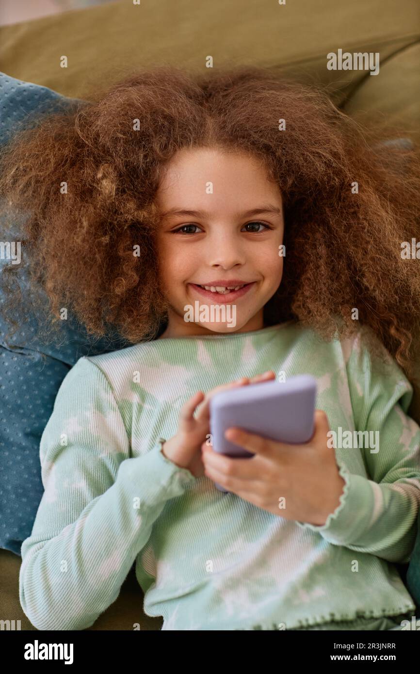 Portrait vertical en haut de la jolie fille regardant l'appareil photo tout en étant couché sur le lit avec un smartphone Banque D'Images