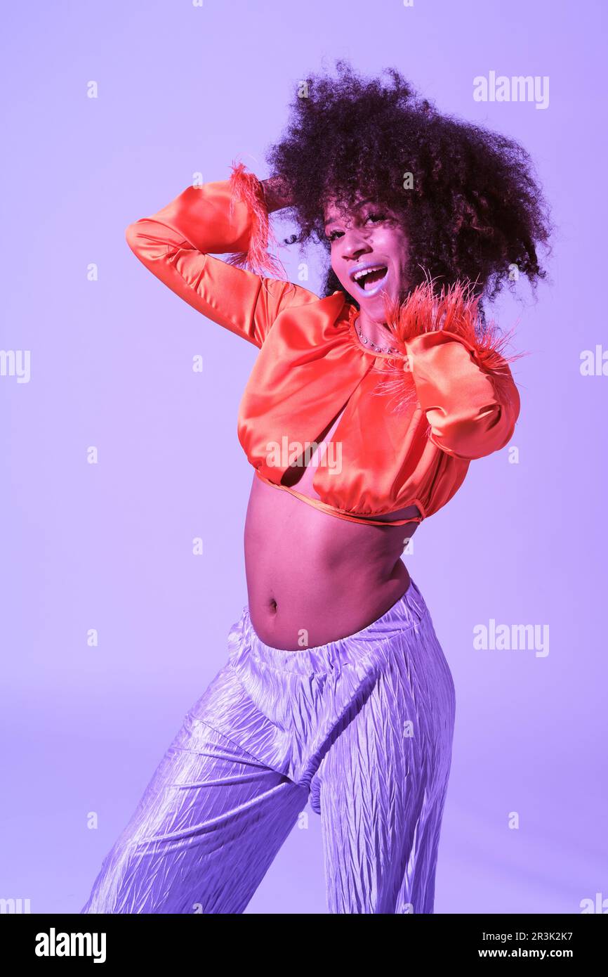 Fille latine avec des cheveux bouclés et des vêtements colorés souriant avec la bouche ouverte et regardant l'appareil photo touchant ses cheveux Banque D'Images