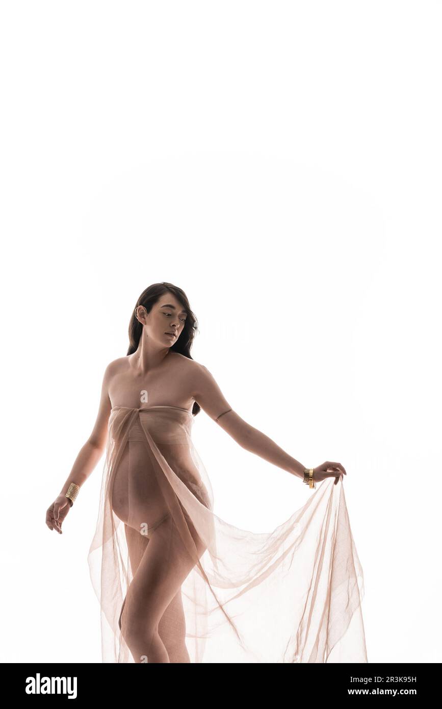 charmante et gracieuse brune femme enceinte posant dans des bracelets dorés et délicate mousseline drapé isolé sur fond blanc, maternité mode Banque D'Images