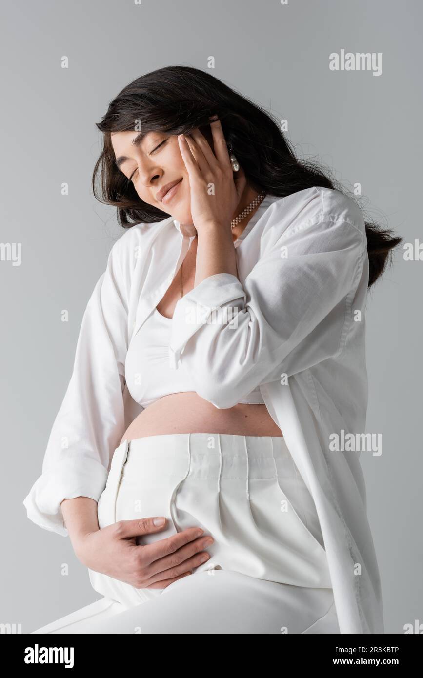 brunette femme enceinte en blanc crop top, chemise et pantalon touchant le ventre et le visage heureux avec les yeux fermés isolés sur fond gris, style de maternité Banque D'Images