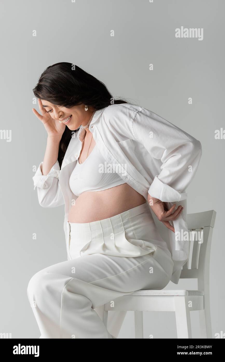 joyeux maman-à-être assis sur la chaise dans blanc petit top, chemise et pantalon tout en tenant la main près du visage heureux isolé sur fond gris, de la mode de maternité Banque D'Images