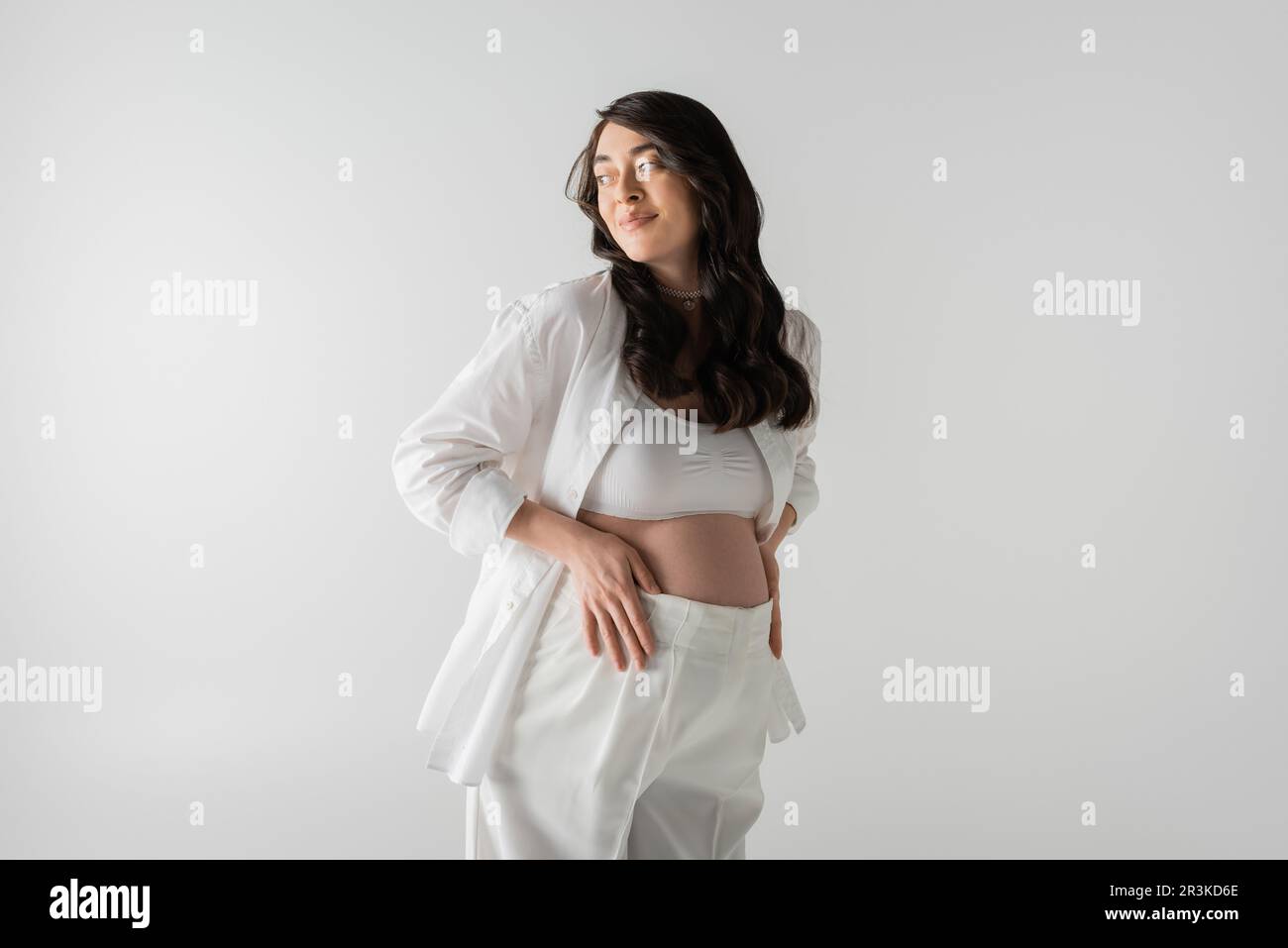femme enceinte gaie en blanc élégant chemise, brassière et pantalon touchant le ventre et regardant loin isolé sur fond gris, maternité conc mode Banque D'Images