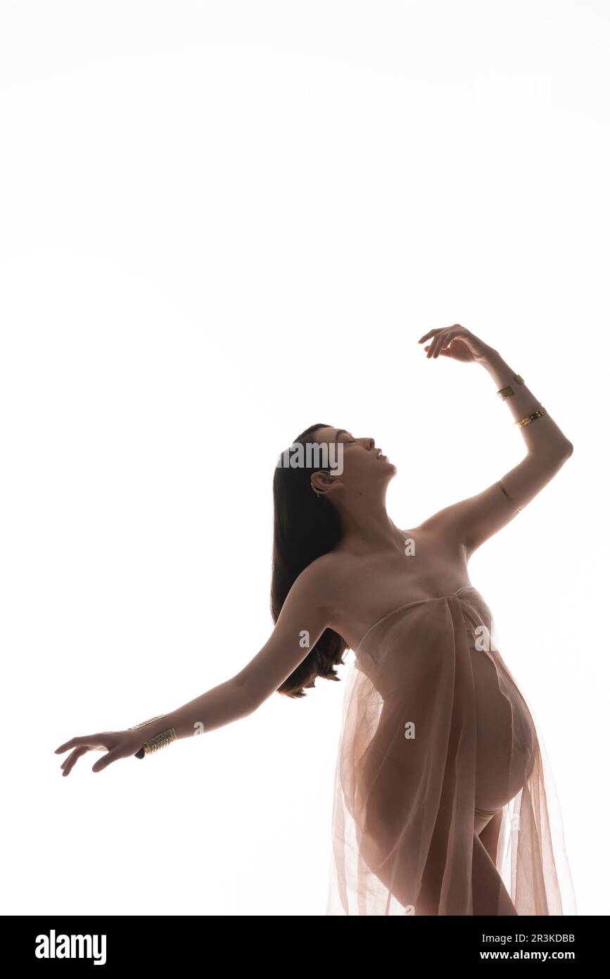 gracieuse mère-à-être dans des bracelets dorés et délicat mousseline drapé posant avec les yeux fermés et la main levée isolée sur fond blanc, maternité Banque D'Images