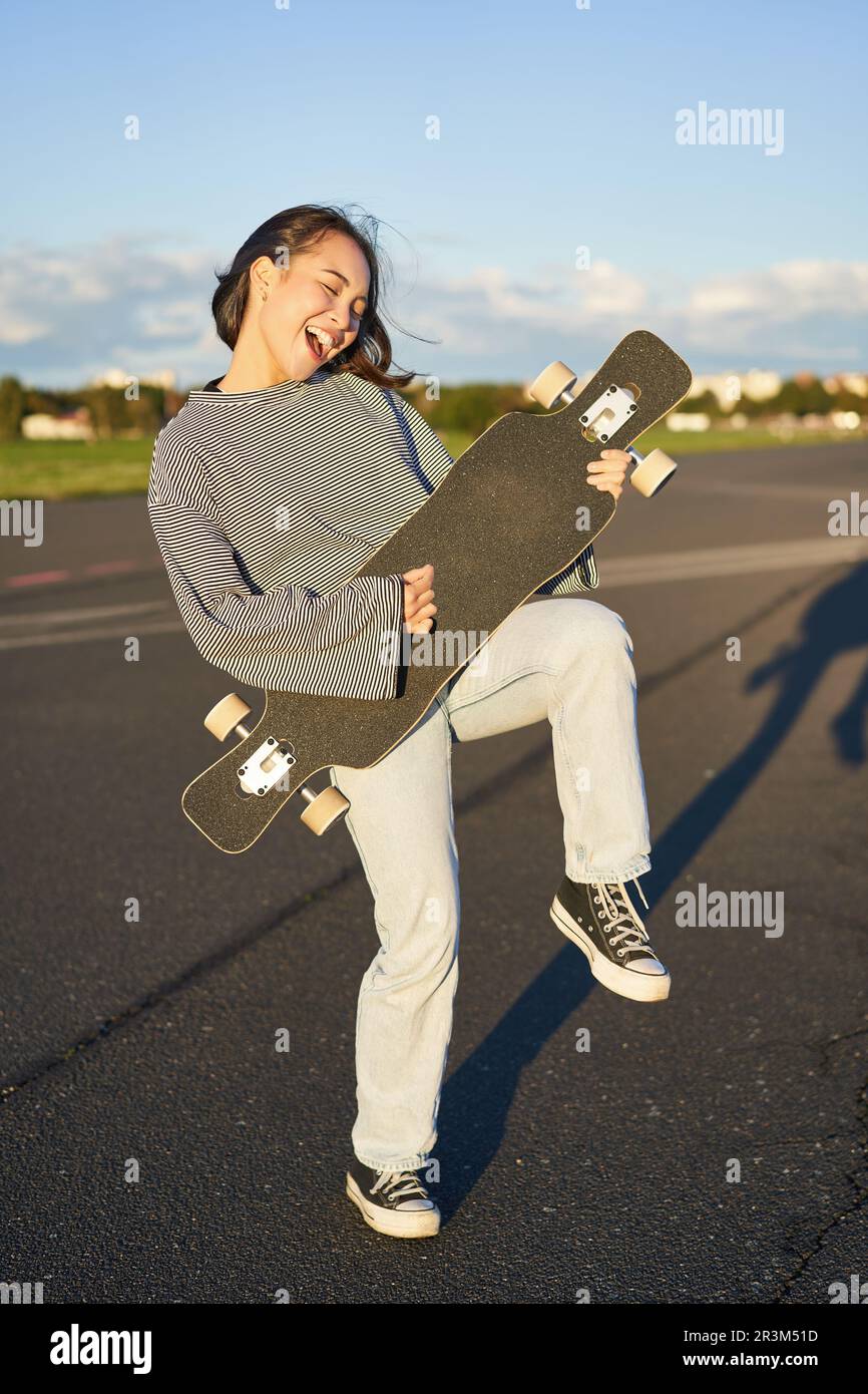 Drôle asiatique fille appréciant le patinage, tenant le skateboard comme la guitare et le jeu d'ombre, ayant l'amusement à l'extérieur Banque D'Images