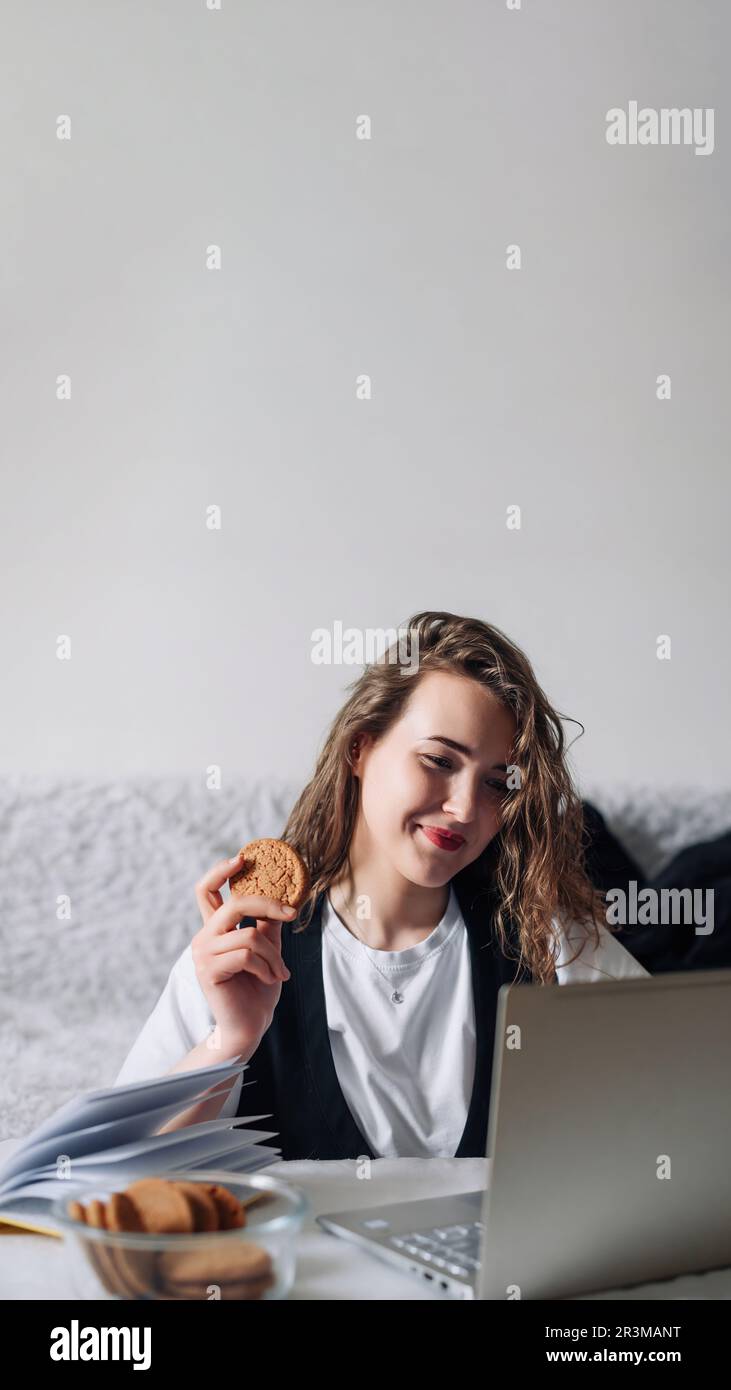 Jeune femme à tête rouge regardant une série ou un podcast regardant l'écran avec intérêt tenant cookie dans la main assis à la table de cuisine avec pot en verre et Banque D'Images