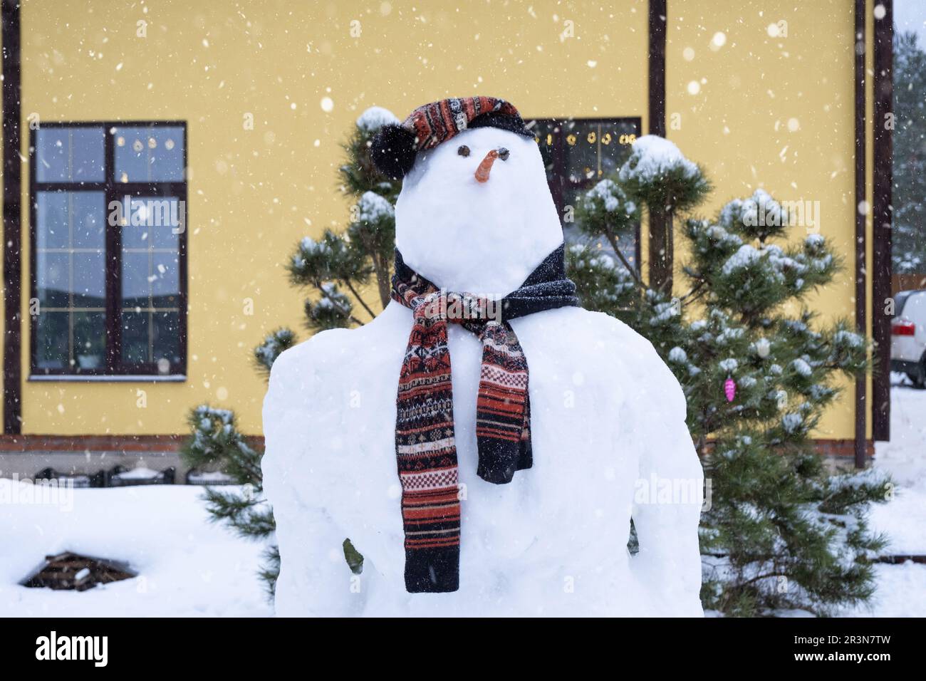 Bonhomme de neige drôle dans un chapeau et un foulard sur le fond d'une maison jaune dans la cour. Hiver, divertissement d'hiver, chute de neige Banque D'Images