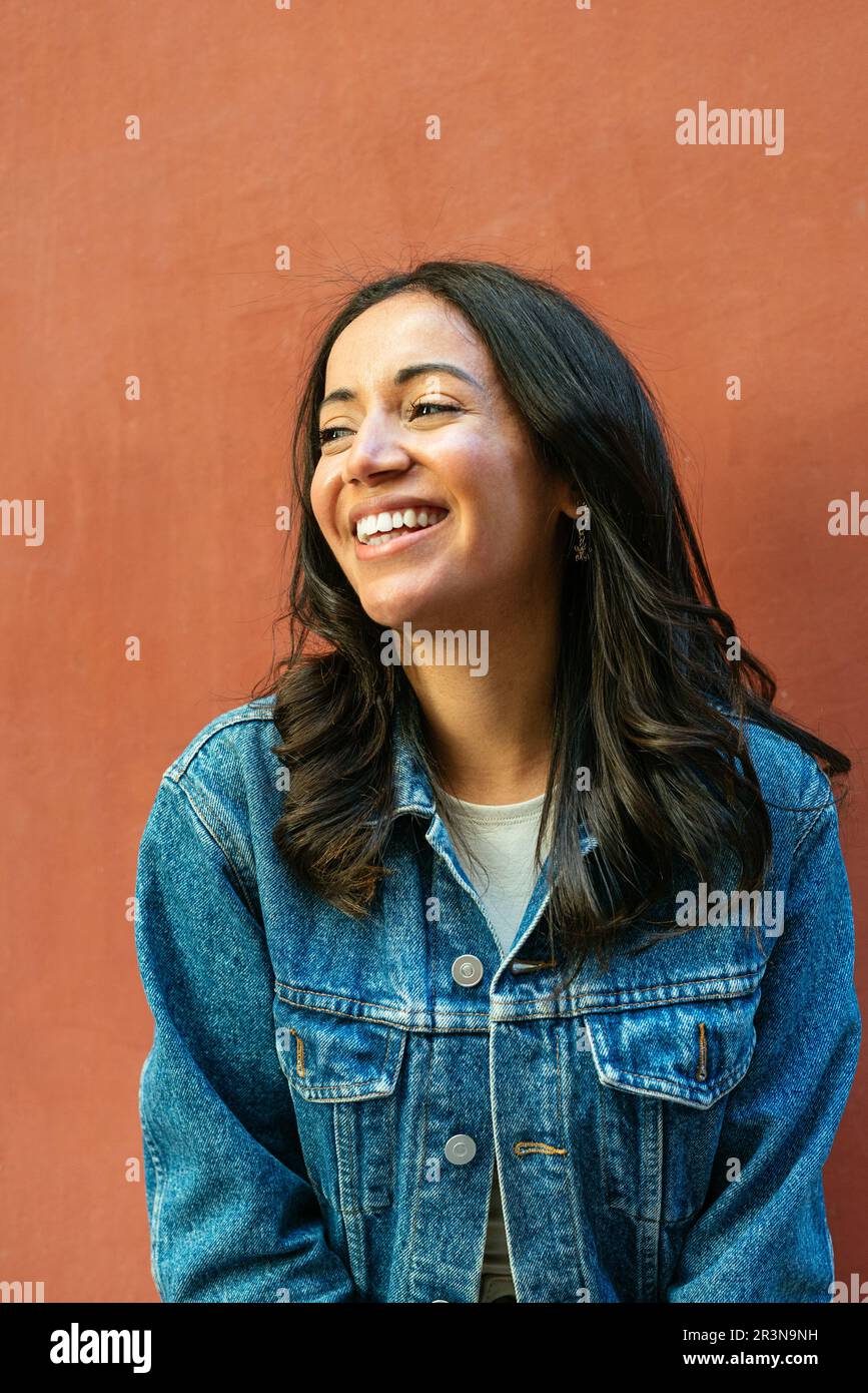Portrait d'une jeune femme hispanique heureuse aux cheveux foncés et en veste en denim souriant tout en regardant loin et debout sur fond orange Banque D'Images