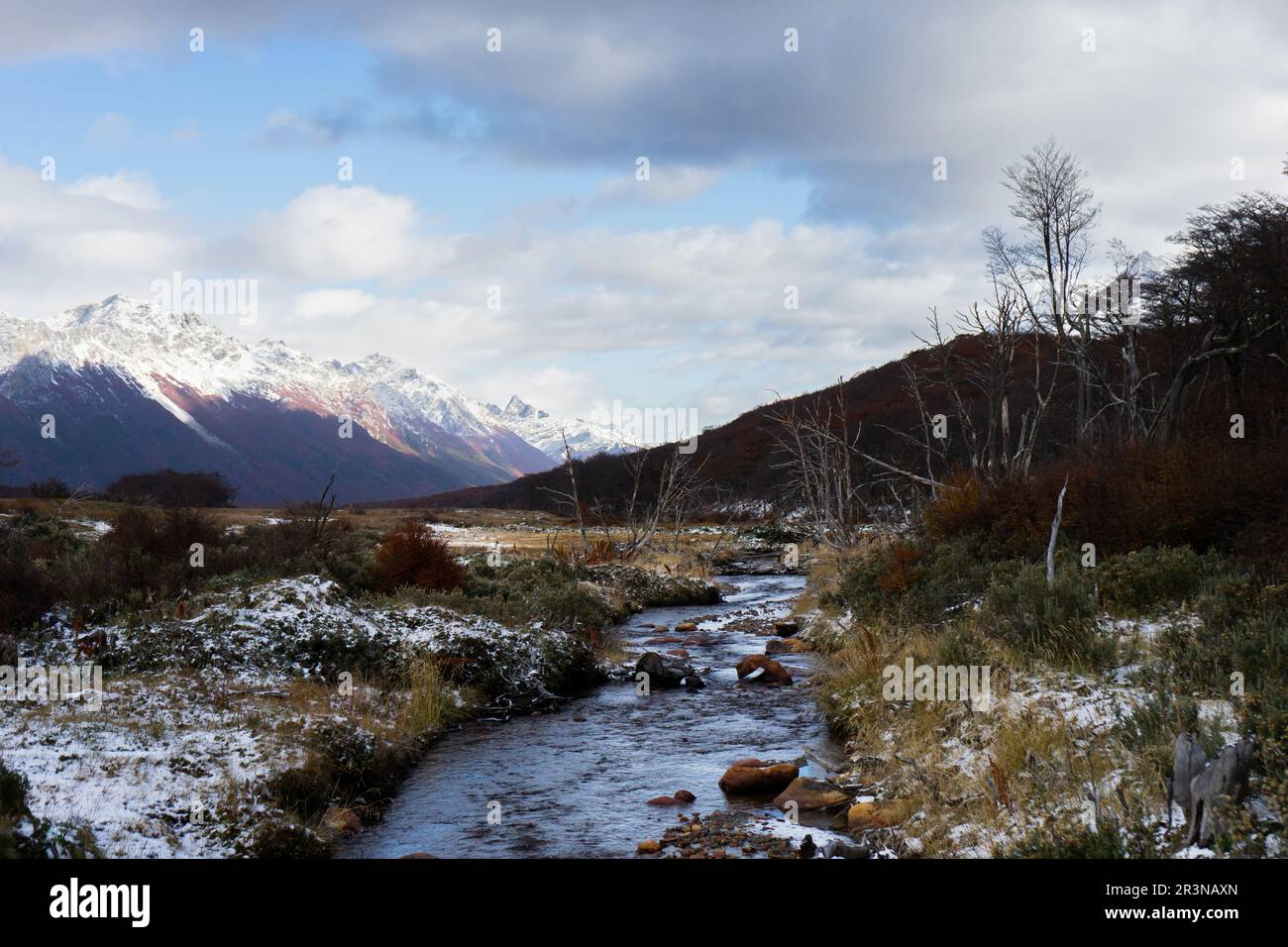 Vue panoramique sur un terrain vallonné avec de l'herbe sèche et du gel sur les côtés de la petite crique avec de l'eau coulante contre des montagnes enneigées et ciel nuageux en winte Banque D'Images