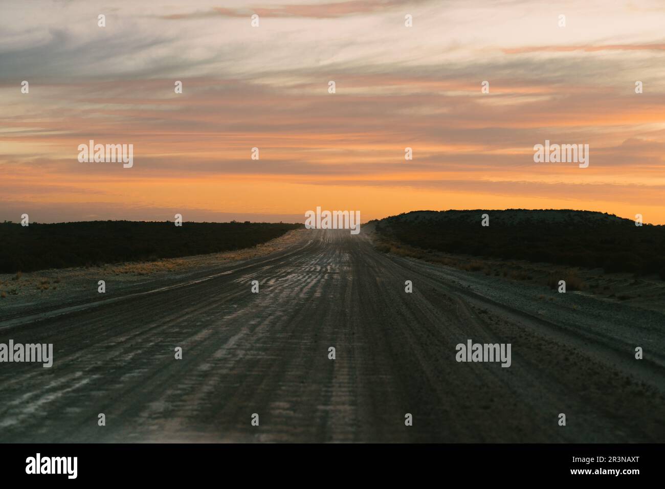 Vue pittoresque d'une route droite vide en construction et de véhicules de transport lourds avec des marques de pneus passant au-dessus d'une colline dans un coucher de soleil nuageux s. Banque D'Images