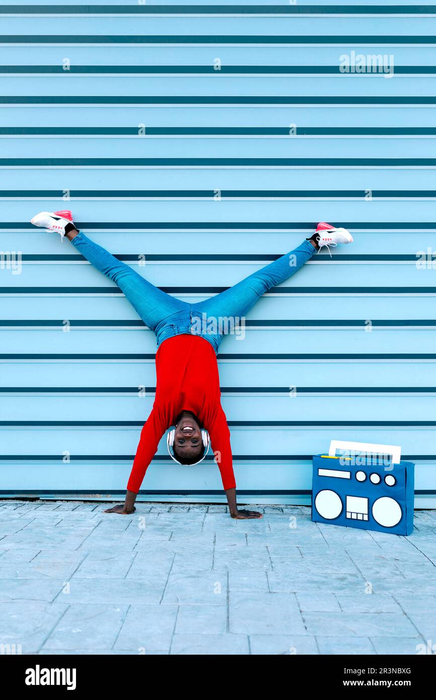 Modèle féminin noir haut de gamme dans un casque qui écoute de la musique et sourit tout en se tenant debout sur les bras à l'envers avec des jambes fendues près de la boombox Banque D'Images