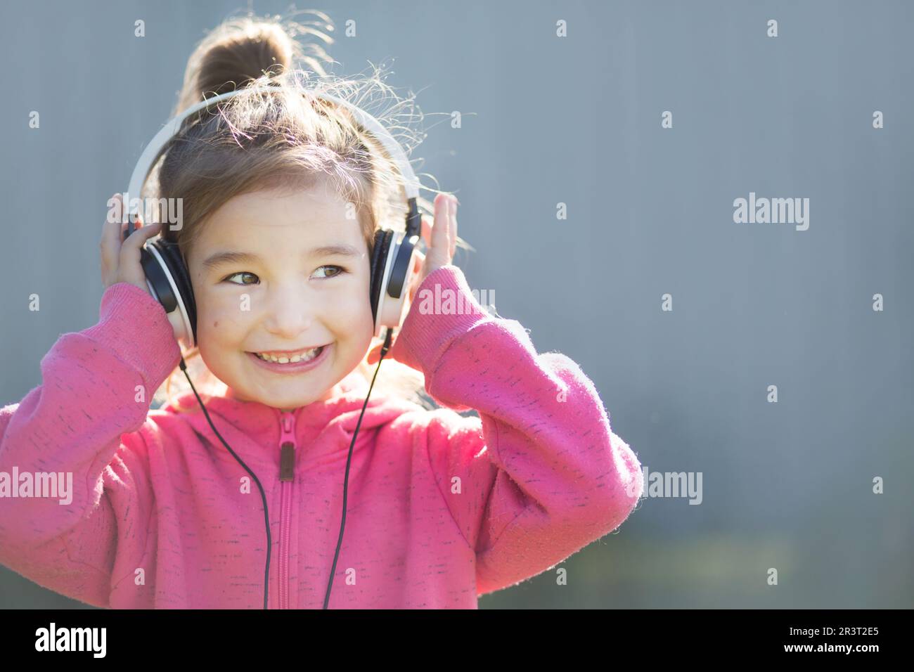 Une jeune fille gaie dans un casque et un pull à capuche rose écoute de la musique et rit. Extérieur, fond gris Banque D'Images