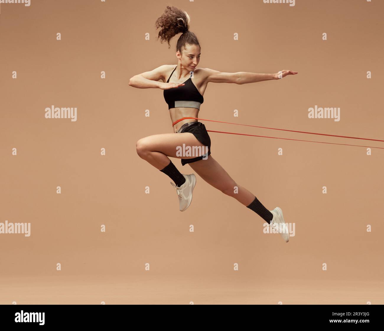 Image dynamique d'entraînement sportif, en forme, forte jeune fille, en mouvement, sautant avec la corde de forme physique sur fond brun clair de studio Banque D'Images