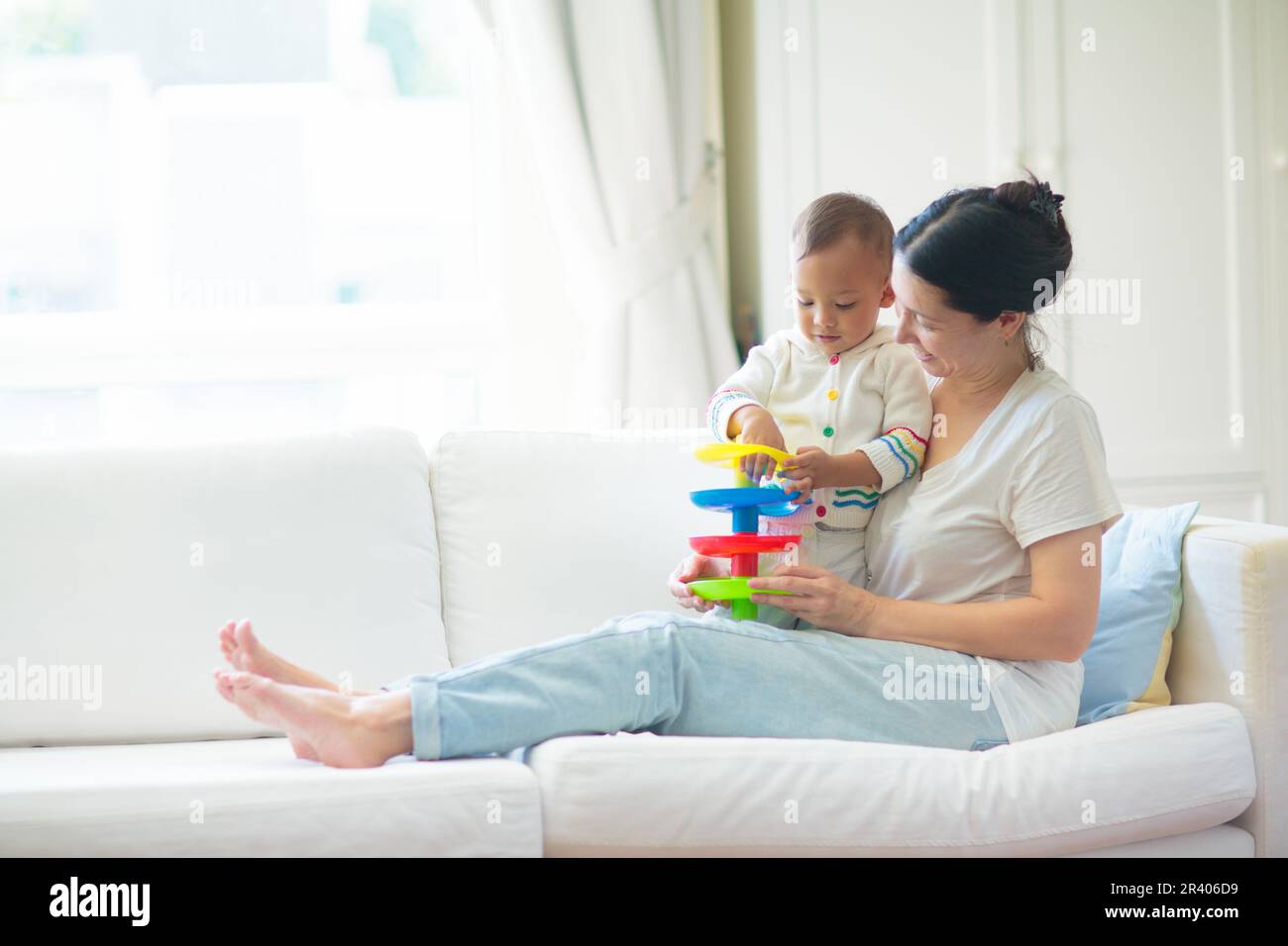 Mère et enfant sur un canapé blanc. Maman asiatique et bébé garçon en couches jouant dans la chambre ensoleillée. Parent et petit enfant se détendant à la maison. Banque D'Images