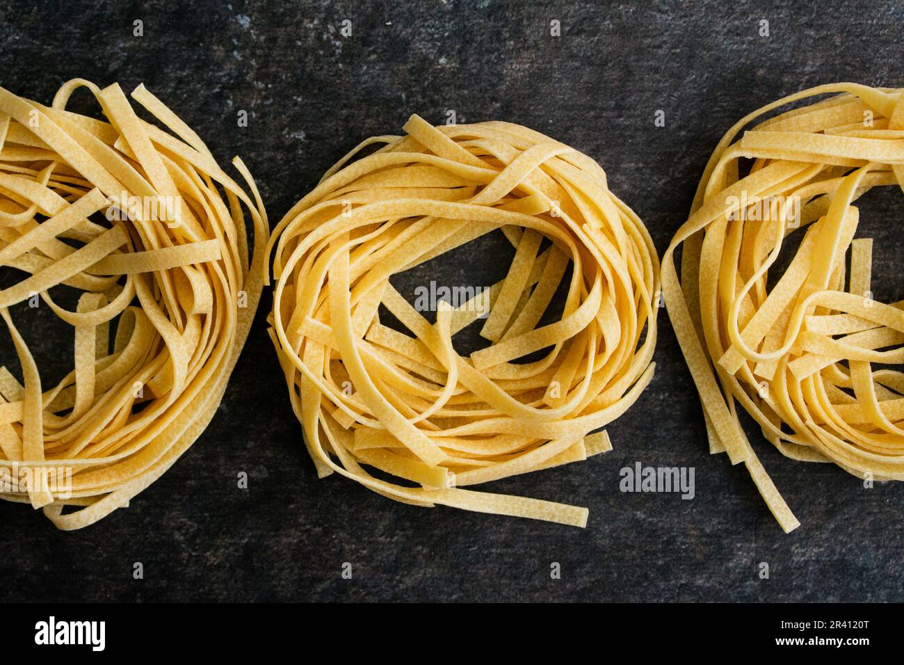 Rangée de trois pâtes Tagliatelle nids sur un fond sombre : nouilles séchées disposées en rangée de nids de pâtes vues directement au-dessus Banque D'Images