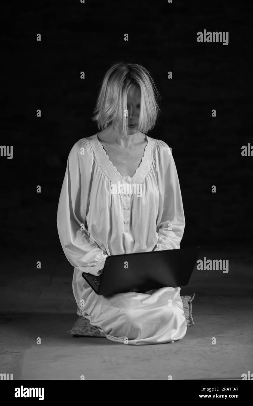 Femme et ordinateur portable dans une pièce sombre vide. Femme d'âge moyen dans une chemise blanche. image en noir et blanc. Banque D'Images