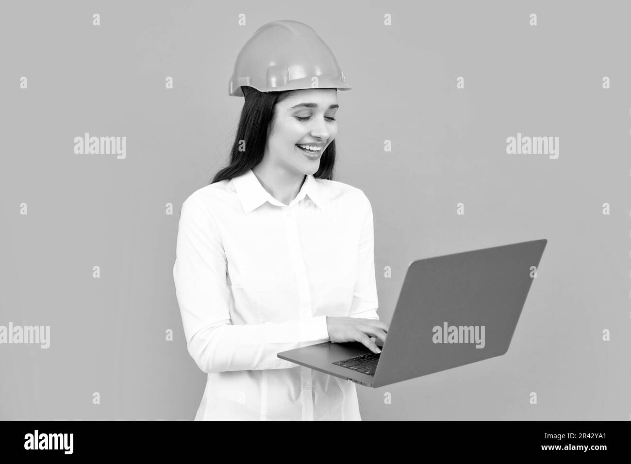Femme souriante positive dans un casque de protection avec ordinateur portable isolé sur fond gris. Jeune femme responsable de la construction. Femme architecte Banque D'Images