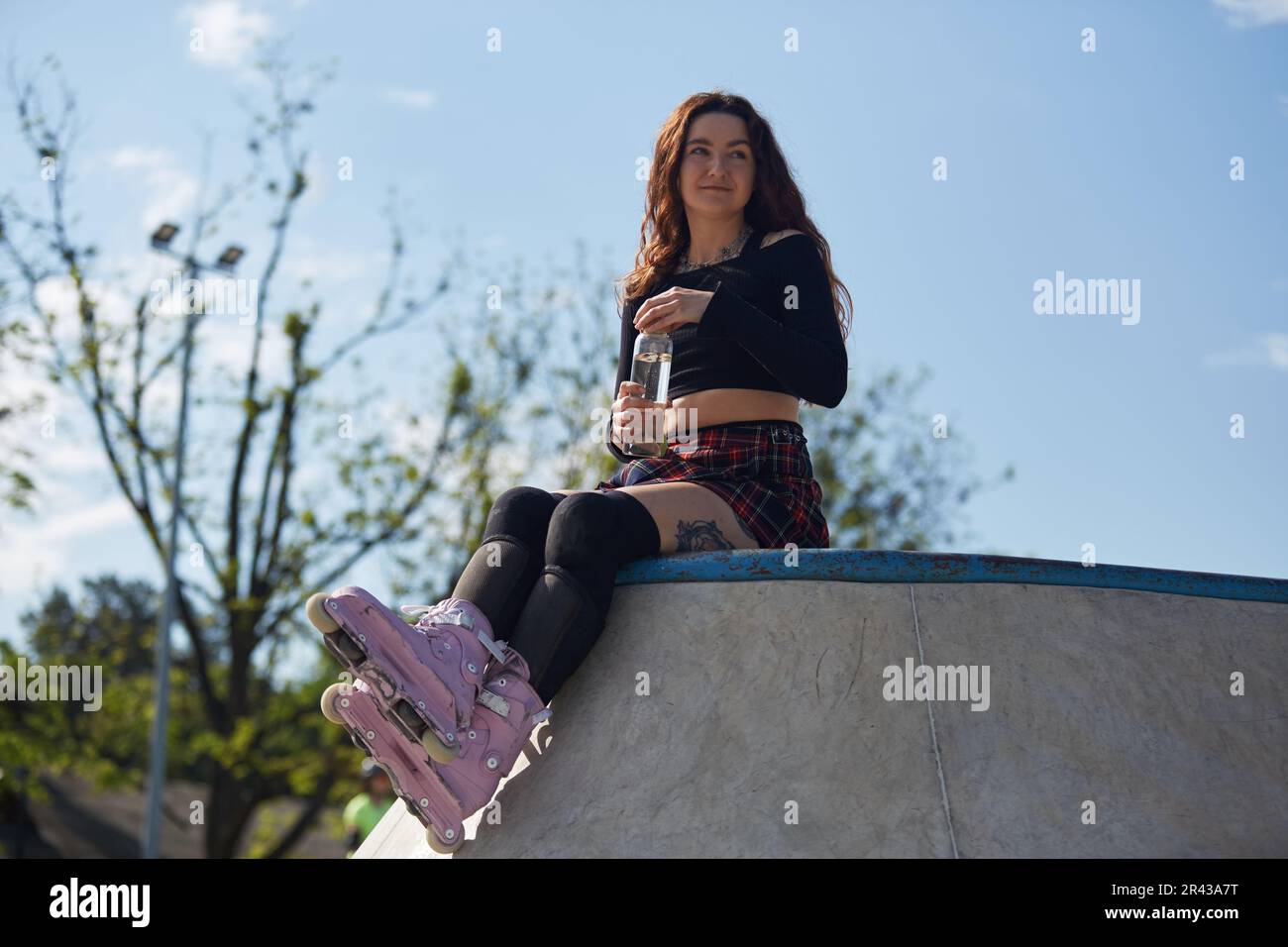 Roller blader femelle assis sur une rampe dans une piscine de patin en béton et de l'eau potable d'une bouteille de verre. Jeune patineuse fraîche portant un agresssiv moderne Banque D'Images