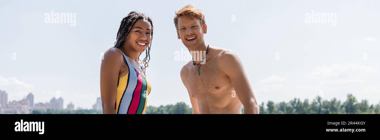 surjoyeuse et sportive redhead homme et attrayante afro-américaine femme en maillot de bain rayé regardant la caméra ensemble sous le ciel bleu extérieur, bannière Banque D'Images