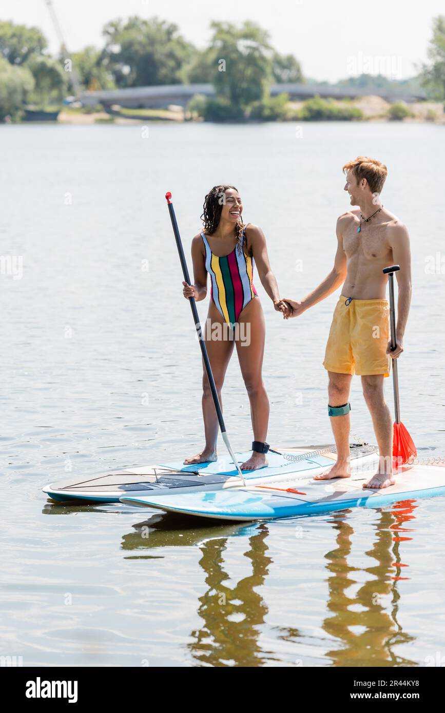 homme à tête rouge actif et femme afro-américaine joyeuse en maillot de bain rayé tenant les mains et souriant l'un à l'autre tout en se tenant debout sur des planches de sup sur lak Banque D'Images