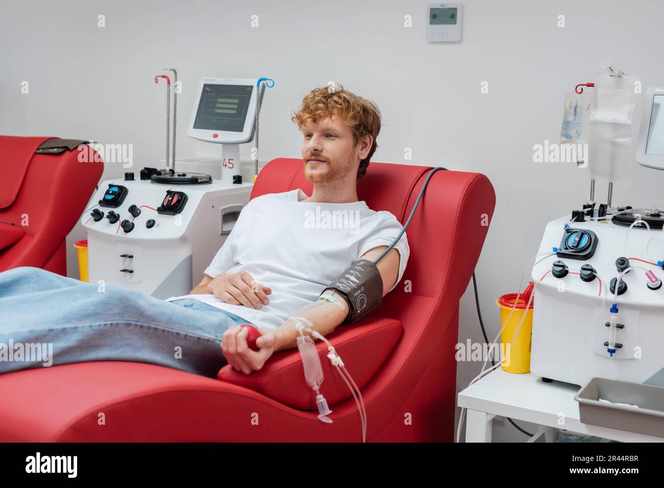 jeune donneur de tête rouge avec brassard de pression et kit de transfusion assis sur une chaise ergonomique et confortable à proximité de l'équipement automatisé et du plast Banque D'Images