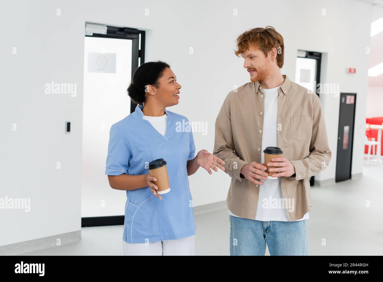 jeune volontaire à tête rouge et travailleur de santé multiracial souriant en uniforme tenant des gobelets en papier tout en parlant dans l'aire d'attente du centre de don de sang Banque D'Images