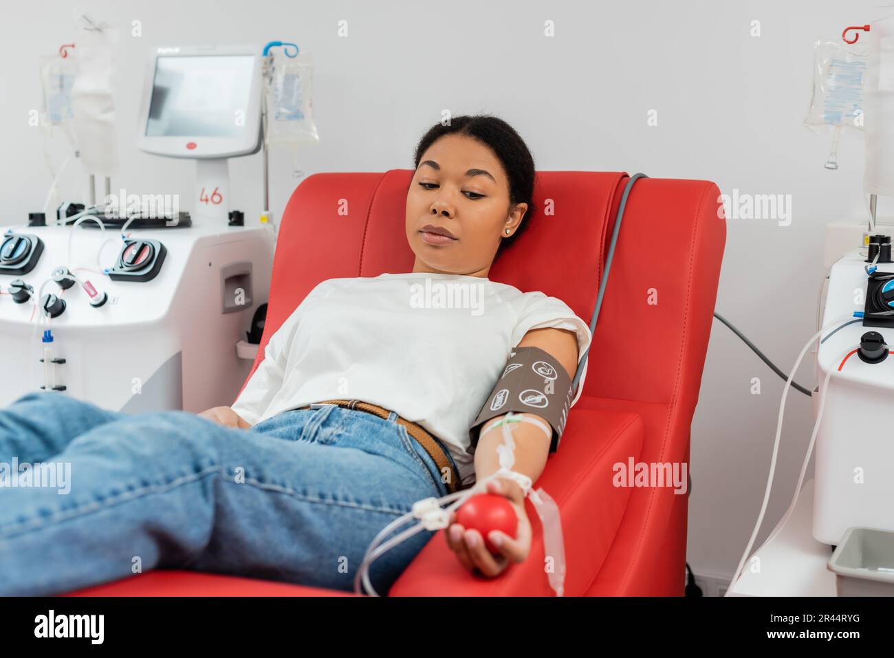 femme multiraciale avec kit de transfusion et boule de caoutchouc assise sur une chaise ergonomique à proximité des transfusions et donnant du sang à l'hôpital Banque D'Images