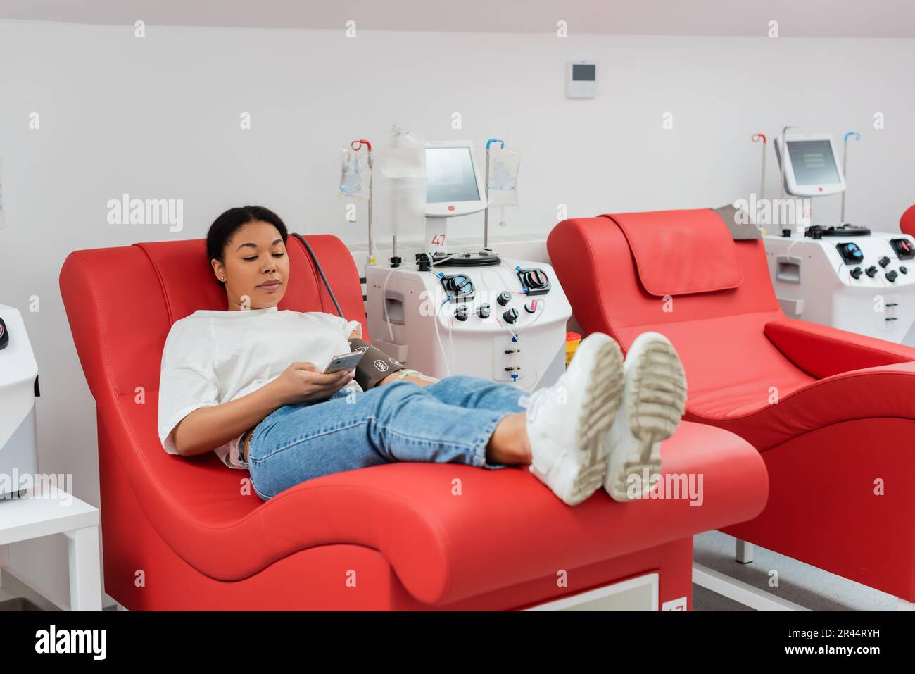 femme multiraciale qui navigue sur internet sur un smartphone tout en étant assise sur une chaise confortable près d'une transfusionnelle tout en donnant du sang dans une clinique, Banque D'Images