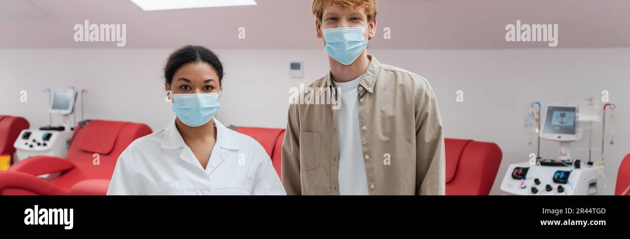 médecin multiracial et volontaire de tête rouge dans les masques médicaux regardant la caméra près des machines de transfusion et des chaises médicales dans le centre de don de sang, ban Banque D'Images