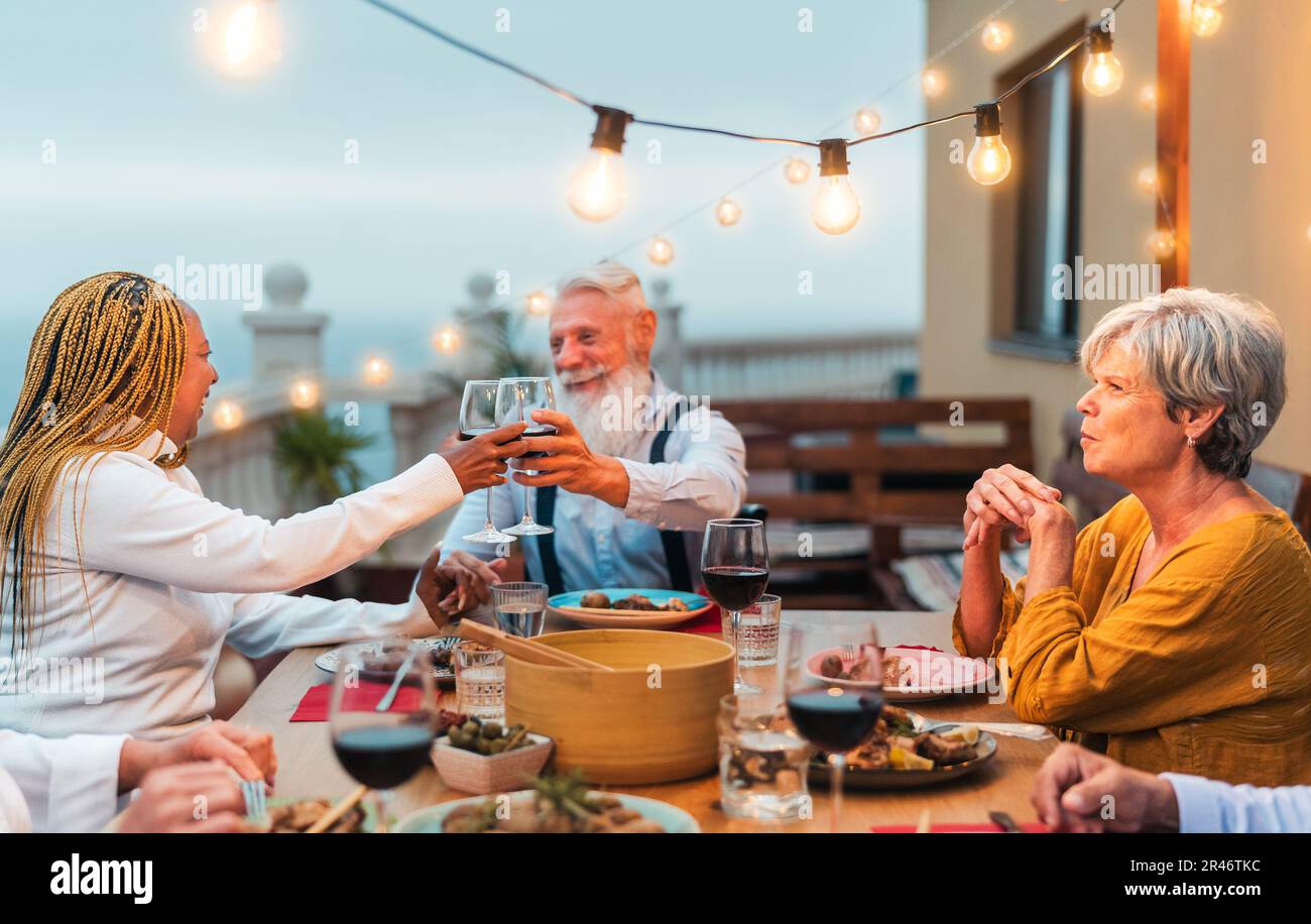 Des amis seniors multiraciaux s'amusent à manger ensemble et à faire du toaster avec Dîner avec vin rouge sur le patio de la maison - repas et vacances concept Banque D'Images