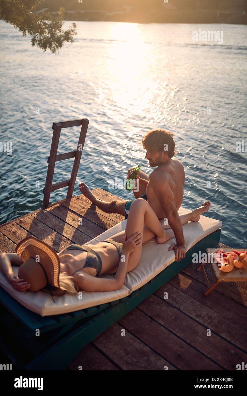 Un jeune couple en maillot de bain bavardant tout en se relaxant et en prenant un bain de soleil sur les rives de la rivière, lors d'une belle journée d'été. Été, rivière, vacances Banque D'Images