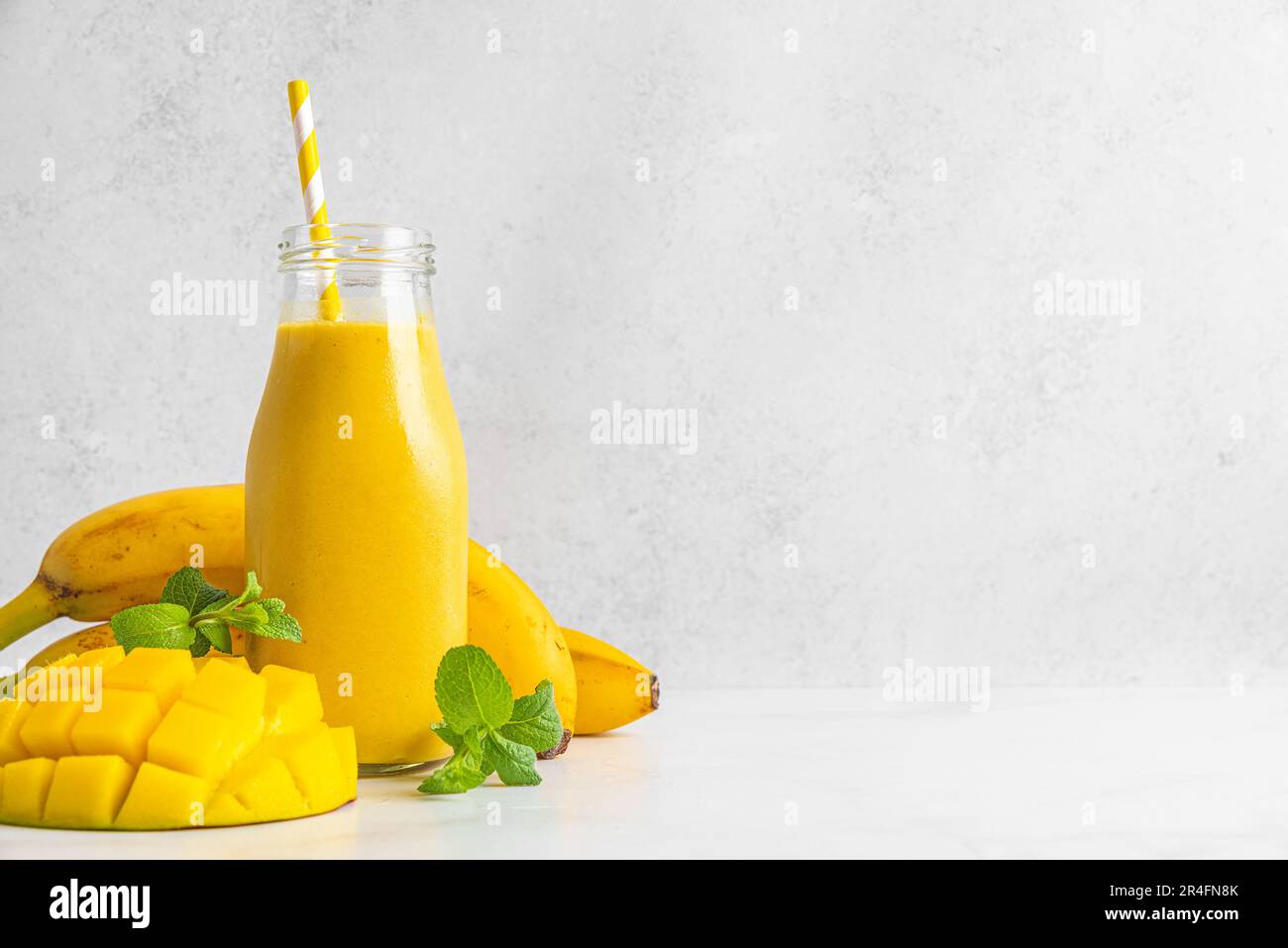 Smoothie rafraîchissant à la mangue tropicale et à la banane dans une bouteille de verre avec fruits frais, menthe et paille sur fond blanc Banque D'Images