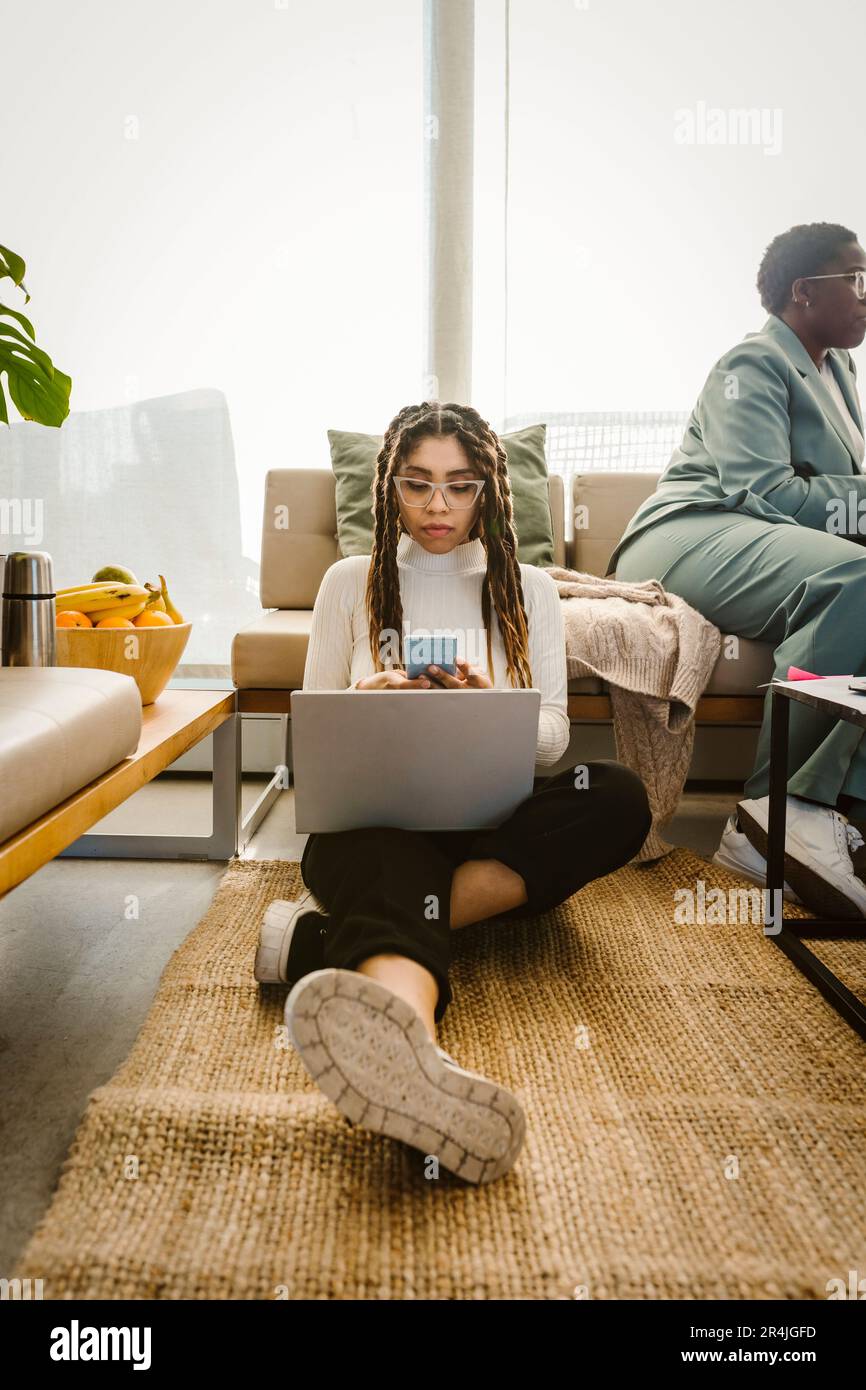 Une femme programmeuse utilisant un smartphone assis avec un ordinateur portable sur un tapis de bureau Banque D'Images