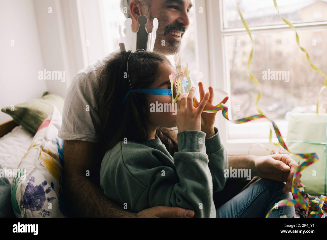 Fille portant une couronne et des lunettes appréciant avec le ruban tout en étant assis sur le père à la maison Banque D'Images