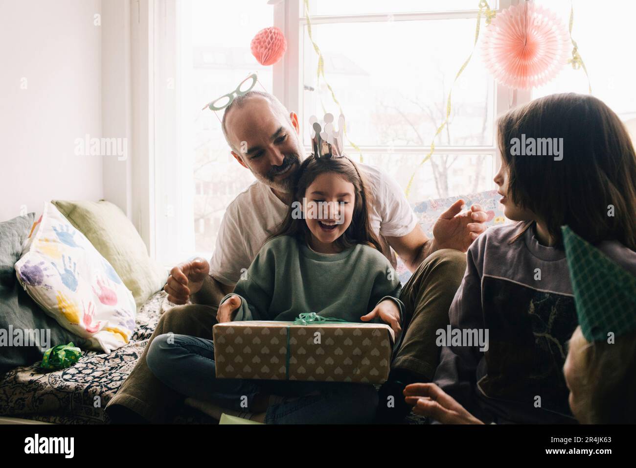 Fille surprise tenant cadeau d'anniversaire tout en étant assise avec frère et père à la maison Banque D'Images
