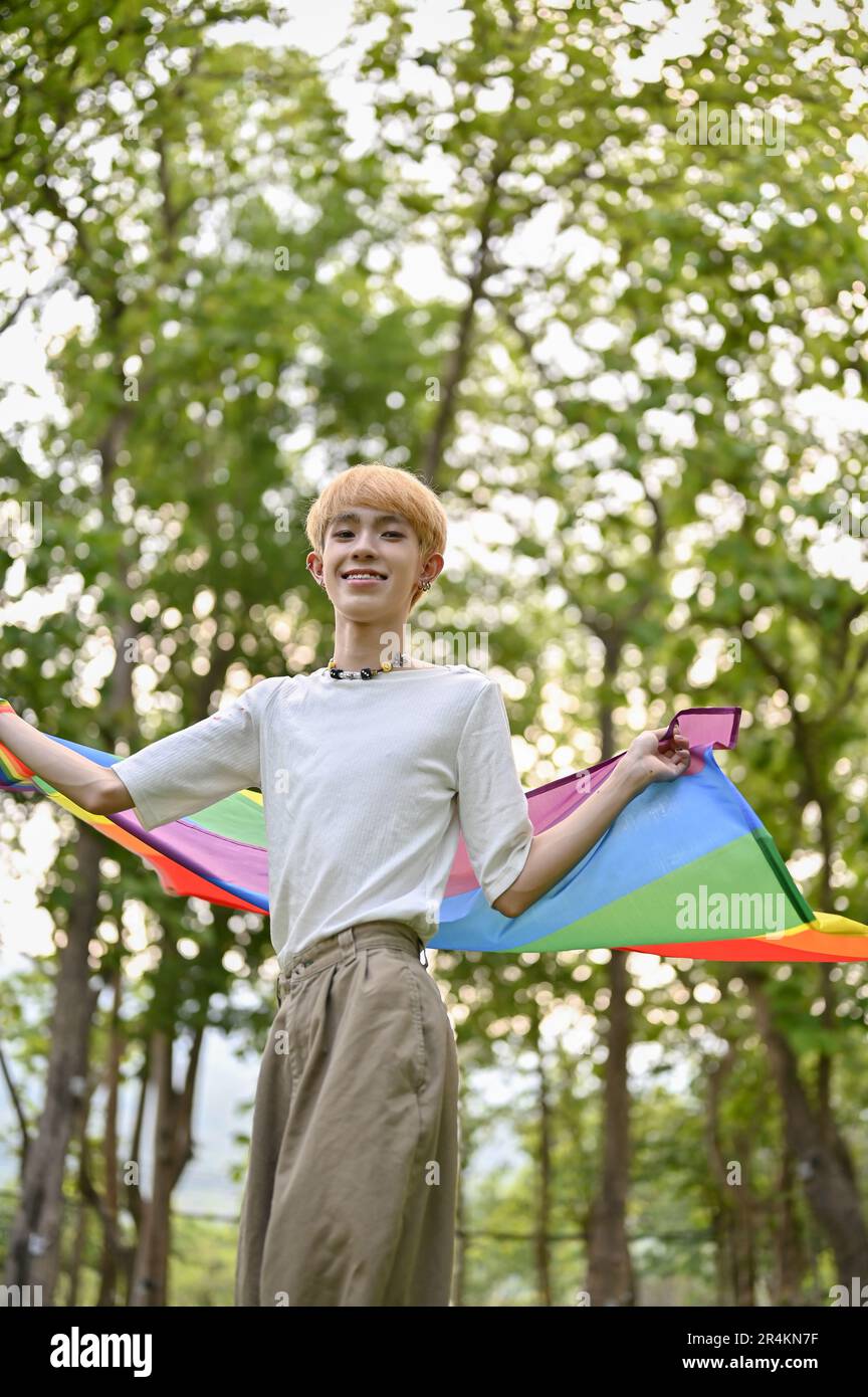 Portrait d'un jeune gay asiatique attrayant et heureux en vêtements décontractés avec un drapeau arc-en-ciel LGBT debout dans le parc. Défilé de fierté, LGBTQ+ Banque D'Images