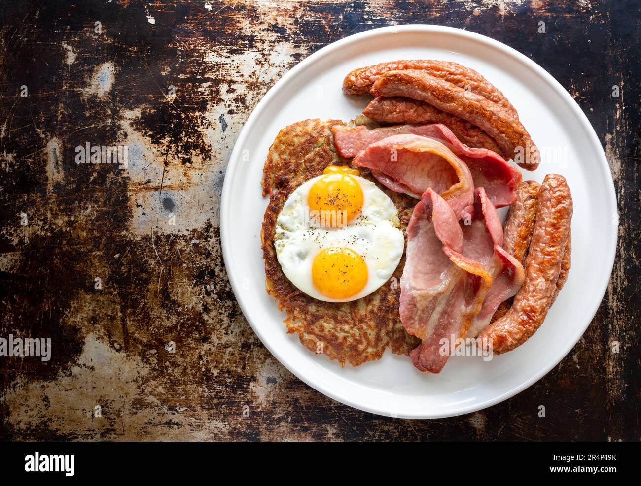 Dégustez un copieux petit déjeuner : saucisse de chipolata, bacon croustillant et œufs frits sur des pommes de terre dorées, servis sur une assiette blanche immaculée Banque D'Images