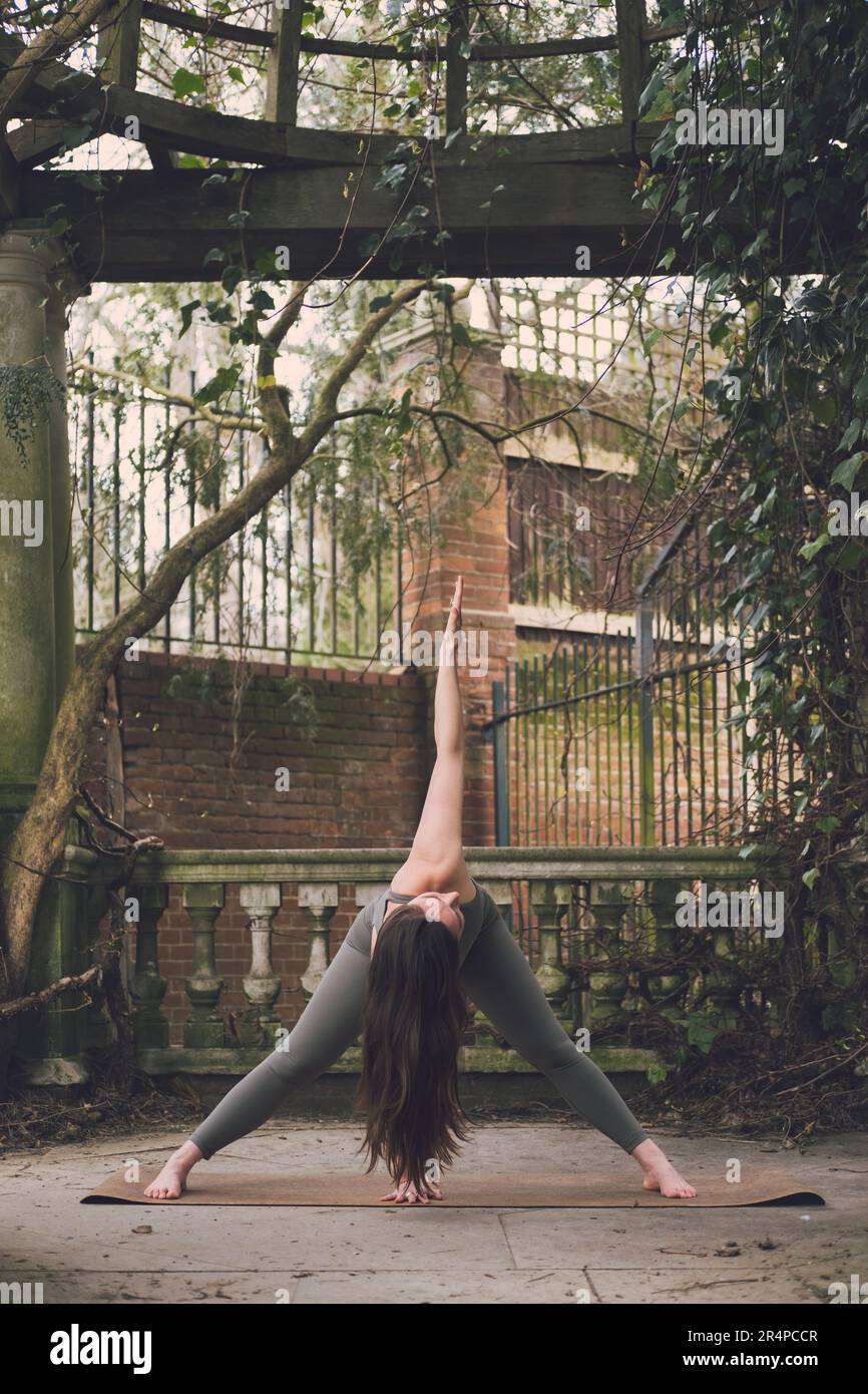 Photo verticale d'un professeur de yoga féminin dans un tour ou Twist, posture du coude en avant à larges pattes dans une terrasse d'un jardin paysagé historique avec des plantes, b Banque D'Images