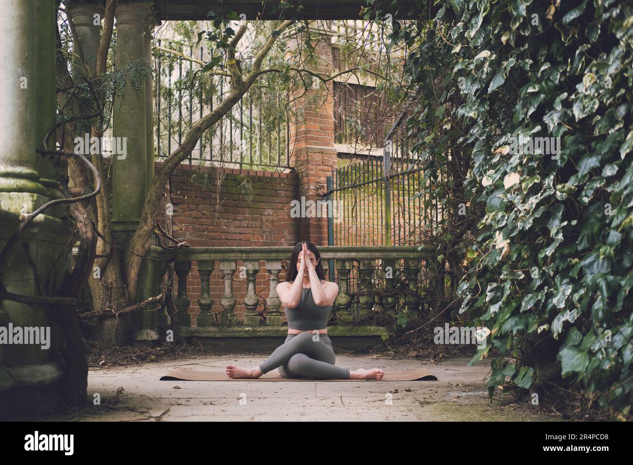 Enseignante de yoga pratiquant dans une terrasse d'un jardin historique avec végétation, colonnes en pierre et balustrade, tenue verte. Gomukhasana (Cow face po Banque D'Images
