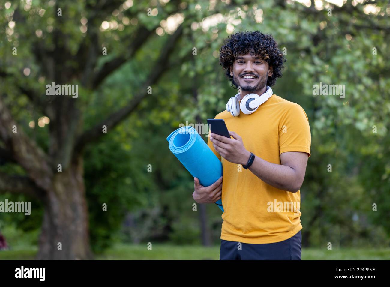 Portrait d'un jeune homme hispanique debout dans un parc, portant un casque, un tapis de maintien et un téléphone. S'est engagé dans des exercices physiques et des sports. Sourire à l'appareil photo. Banque D'Images