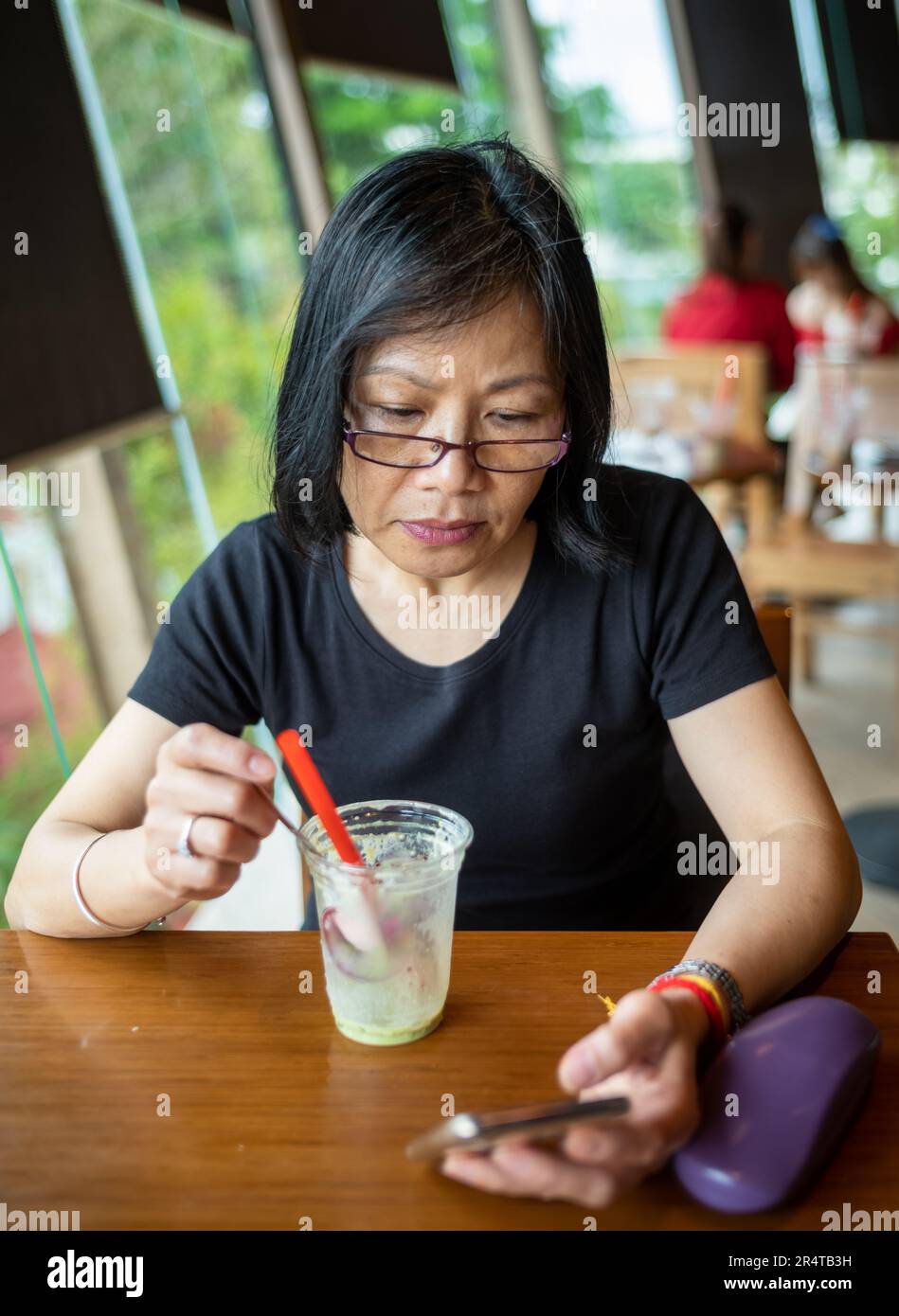 Une femme vietnamienne d'âge moyen termine son café comme elle lit sur son smartphone, Danang, Vietnam. Banque D'Images