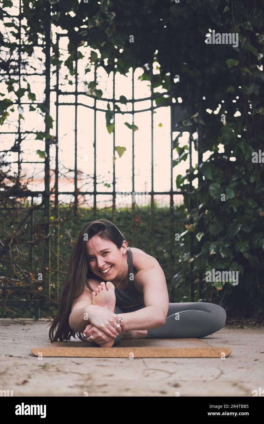 Femme heureuse professeur de yoga dans une jambe plié en avant Bend pose regardant l'appareil photo sourire, cheveux longs brunette, tenue verte, tapis de liège, extérieur, plante un Banque D'Images